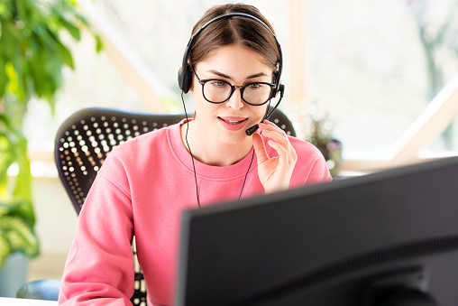 Young businesswoman in headset speaking by conference call while looking at computer. Home office.