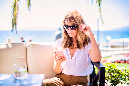 Portrait shot of attractive mature woman sitting on lounge sofa by the seaside while relaxing outdoor and text messaging.
