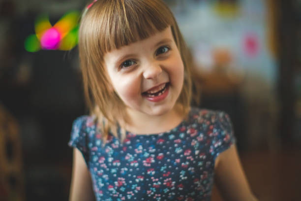 4 years girl joking, sticking tongue out with silly expression. Child making face. Funny foolishness portrait of little kid doing crazy comical gesture among a white wall. Closeup face. Low contrast. Adorable Caucasian kid doing crazy  gesture at home in bedroom. Messing around, grimacing.  Naughty, play pranks, misbehave