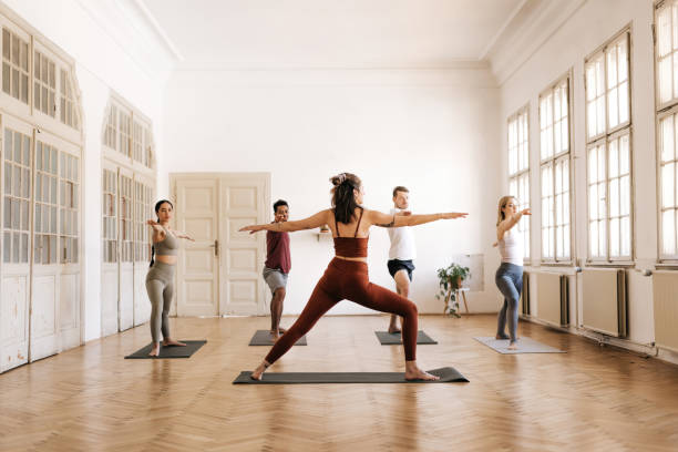 Group of people in Warrior 2 Pose in a bright yoga studio A yoga instructor is teaching the Warrior 2 pose in a spatious bright room with large windows. They are dressed in trendy sports clothing. Horizontal daylight photo. yoga instructor stock pictures, royalty-free photos & images