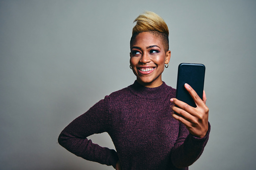 Smiling young female hipster talking selfie through smart phone against gray background