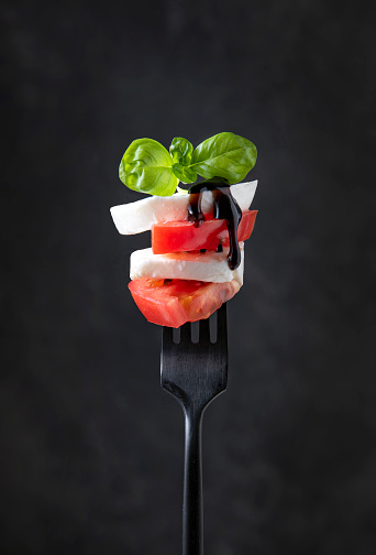 Caprese salad. mozzarella slices, fresh tomatoes and fresh basil leaves with balsamic sauce on a black fork on a dark background. Front view
