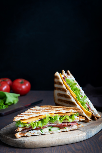 Simple tasty sandwich consisting of cucumber, slice of meat and cheese, lettuce and tomatoes between slices of grilled bread served on cutting board on dark brown wooden table at kitchen. Vertical
