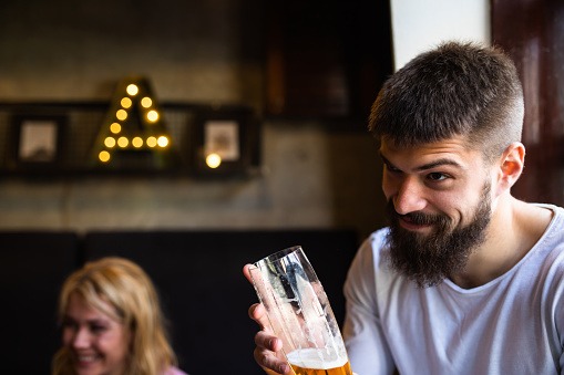 A young man is raising his glass and making a funny face while out for drinks with friends