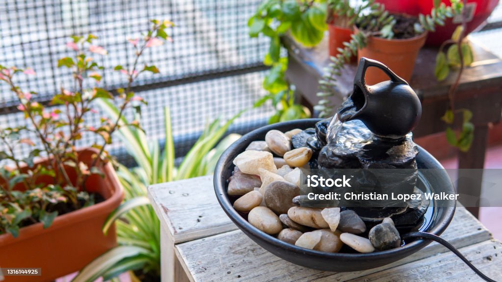 zen water fountain Close view of a black  decorative zen water fountain on top of a small white wooden table next to a few green plants on a balcony. Taken on a warm summer morning with natural lighting Decoration Stock Photo