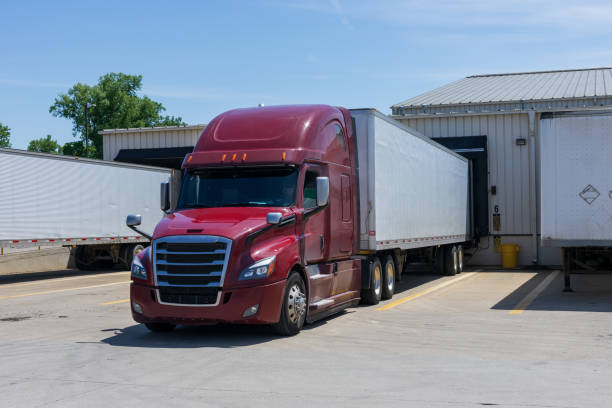 Modern red big rig semi truck with a high cabin transporting a dry van semi trailer with commercial cargo are loading  at a warehouse. Modern red big rig semi truck with a high cabin transporting a dry van semi trailer with commercial cargo are loading  at a warehouse in an industrial park. A truck driver is in the cab. All logos removed. loading bay stock pictures, royalty-free photos & images