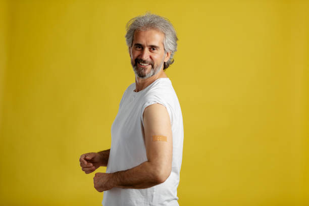 Happy vaccinated mature man showing shoulder with plaster bandage after Covid-19 vaccine injection. stock photo