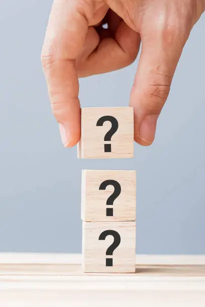 Photo of hand holding Questions Mark ( ? ) on wooden cube block on table background. FAQ( frequency asked questions), Answer, Q and A, Information, Communication and interrogation Concepts