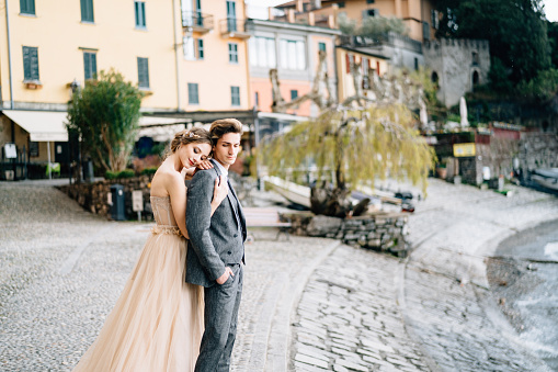 Bride hugs groom from behind, resting her head on his shoulder, standing on the embankment of Lake Como against the background of old houses. High quality photo