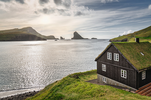Faeroe Islands Drangarnir Rocks and Tindholmur on the North Atlantic Ocean Horizon. Seascape View in warm afternoon sunset light. Typical faroese grass roof house in the foreground at the Water`s Edge towards the North Atlantic Ocean. Faroe Islands, Vágar Island, Kingdom of Denmark, Nordic Countries, Europe