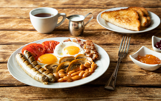Traditional morning breakfast in England - sausages, tomatoes, eggs, bacon, mushrooms, white beans, toasted toast, a cup of black coffee and jam on a wooden table -  side view and close-up