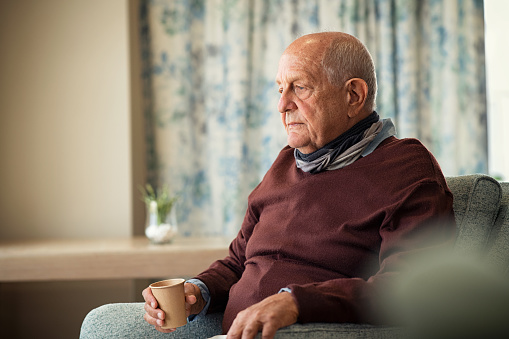 Depressed senior man sitting on armchair holding disposable cup of coffee and thinking. Frustrated retired man sitting on sofa and drinking a cup of hot tea. Sad mature man sitting alone at nursing home with sad expression.