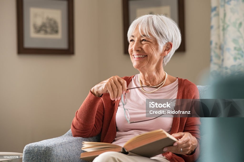Happy senior woman smiling at home Cheerful senior woman holding book and eyeglasses thinking while relaxing at home. Happy elderly woman reading book at home sitting on couch. Beautiful old teacher takes a break from reading while looking through the window with a big grin. Senior Adult Stock Photo