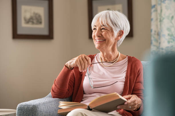 Happy senior woman smiling at home Cheerful senior woman holding book and eyeglasses thinking while relaxing at home. Happy elderly woman reading book at home sitting on couch. Beautiful old teacher takes a break from reading while looking through the window with a big grin. old stock pictures, royalty-free photos & images