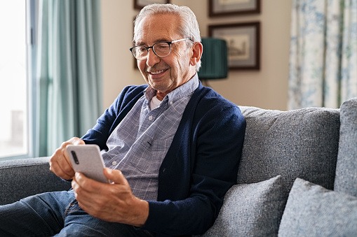 Smiling old man using smartphone while sitting on couch in living room. Happy elderly man with spectacles messaging on mobile phone at home. Healthy senior in video call with his family on smart phone while relaxing at nursing home.