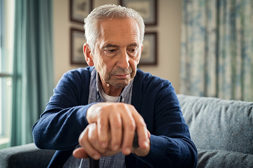 Depressed old man sitting at home while holding walking stick. Retired sad man holding wooden walking cane handle sitting alone at care facility. Elderly man suffering from loneliness and alzheimer at nursing home.