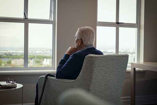 Rear view of senior man sitting on armchair and looking through the window. Lonely old man sitting at home near window with walking cane during covid19 outbreak. Thoughtful retired man abandoned at nursing home facility.