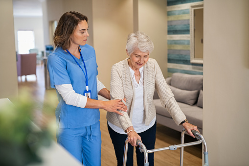Lovely nurse helping old woman to walk at nursing home with walker. Young nurse helping senior patient using a walking frame to walk in hospital corridor. Friendly caregiver and disabled lady in care facility after rehabilitation session.