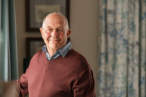 Portrait of happy retired senior man standing at home near window. Satisfied old man in casual clothes looking at camera and smiling while standing near the window. Positive and confident elderly enjoy his retirement at care facility.