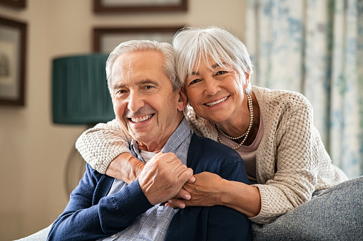 Portrait of romantic senior man with his beautiful wife stay at home. Smiling and caring old woman embracing from behind her retired husband sitting on couch. Cheerful old couple looking at camera with joy.