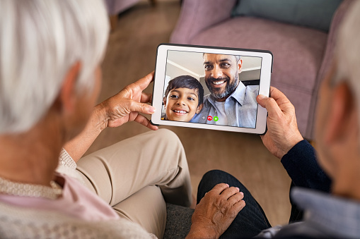 Rear view of senior couple doing a video call with son and grandson during covid19 pandemic. Close up of hands of old grandparents holding digital tablet while in conversation with little grandson. Happy indian father with child greeting in video conference his parents.