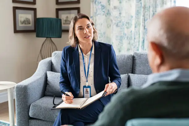 Mature social worker talking to senior man and taking notes of health progress. Mid adult woman in formal clothing visiting old man at home for medical history. Female support worker talking to elder in living room.