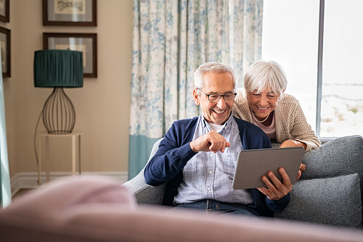 Happy old couple sitting on sofa using digital tablet with copy space. Cheerful senior man and beautiful elderly woman relaxing at home while watching funny video on digital tablet. Smiling grandparents making a video call with their nephews during lockdown.