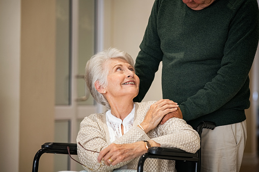 Senior husband comforting woman on a wheelchair by holding hand and looking at her at nursing home. Happy disabled woman at facility care center with her husband. Old man pushing handicapped woman sitting on a wheelchair in the corridors of the medical clinic.