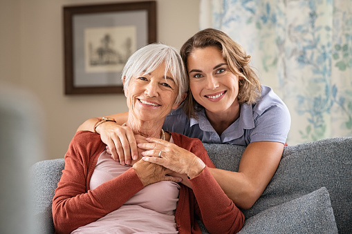 Cheerful mature woman embracing senior mother at home and looking at camera. Portrait of elderly mother and middle aged daughter smiling together. Happy loving daughter hugging from behind elderly mom.