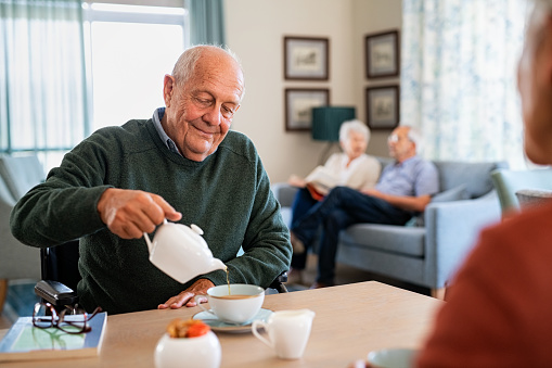 Smiling old man pouring hot tea in cup from kettle in nursing home. Elderly man in care centre sitting on wheelchair and drinking tea. Happy disabled elder in wheelchair enjoying tea time in the afternoon together with other patient of the care facility.