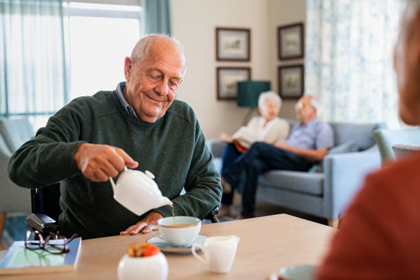 Serene senior man enjoy tea time at nursing home Smiling old man pouring hot tea in cup from kettle in nursing home. Elderly man in care centre sitting on wheelchair and drinking tea. Happy disabled elder in wheelchair enjoying tea time in the afternoon together with other patient of the care facility. community center food stock pictures, royalty-free photos & images