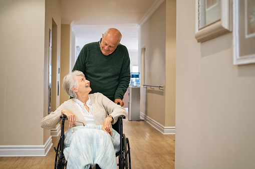 Old man pushing disabled wife on wheelchair in hospital hallway. Happy smiling senior woman in a wheelchair relaxing and walking with her husband in care centre during a visit. Elderly man pushing his old wife after a physiotherapy session or medical exam.
