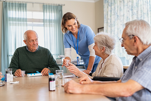 Young nurse in uniform giving medicine to group of seniors at retirement community. Happy smiling nurse gives medicine to elderly patients during her shift in a nursing home. Happy senior woman taking her dose of medicines from helpful caregiver at hospice.