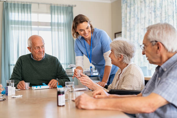 Lovely nurse giving medicine to senior woman at care facility Young nurse in uniform giving medicine to group of seniors at retirement community. Happy smiling nurse gives medicine to elderly patients during her shift in a nursing home. Happy senior woman taking her dose of medicines from helpful caregiver at hospice. nursing home stock pictures, royalty-free photos & images