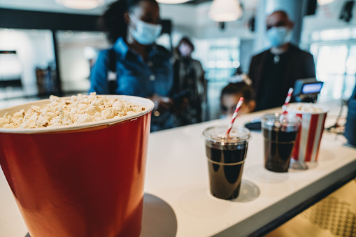 Focus on popcorn and drinks at the bar counter. Movie theater's lobby. Defocused people waiting in line in the background.
