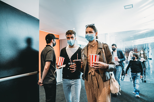 Millennial couple is entering in the cinema hall together, holding popcorn and drinks. They are wearing protective face masks.