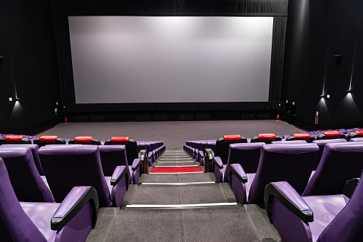 Empty big movie theater hall with a white screen for copy space. Wide angle view of the purple seats and the room, looking at the screen.