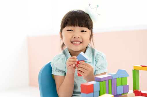 Cute little Asian girl playing with colorful toy blocks while sitting at table and smiling at the camera. Educational and creative toys and games for young children. Child at home.