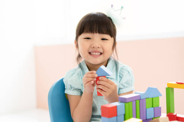 Cute little Asian girl playing with colorful toy blocks while sitting at table and smiling at the camera. Educational and creative toys and games for young children. Child at home. Cute little Asian girl playing with colorful toy blocks while sitting at table and smiling at the camera. Educational and creative toys and games for young children. Child at home. asian childcare centre stock pictures, royalty-free photos & images