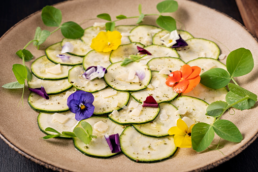 Recipe of; zucchini sliced  with a mandolin, lightly marinated with a pesto and lemon  juice marinade, seasoned with salt, pepper, and vegan parmesan cheese and decorated with edible pansy flowers and pea shoots. Colour, horizontal format.