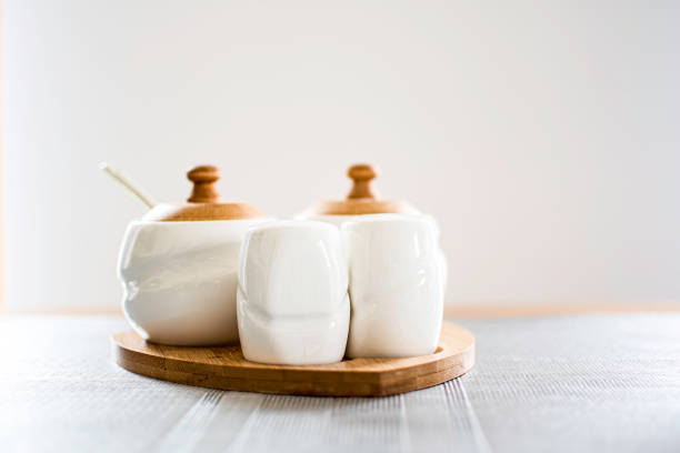 White seasoning containers on the wood. Modern design kitchen detail. Salt, pepper and sugar containers made from ceramics. Selective focus on the small container in front, blurred background.
