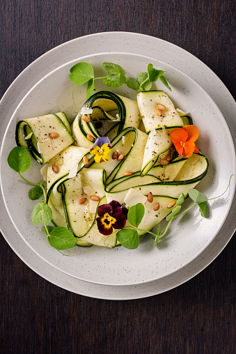 Recipe of; zucchini sliced length ways with a mandolin, lightly marinated with a pesto and lemon  juice marinade, seasoned with salt, pepper, pine nuts and vegan parmesan cheese and decorated with pea shoots and edible flowers. Colour, vertical format.