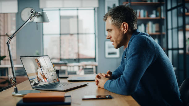 istock Handsome Caucasian Man Having a Video Call with Male Doctor on Laptop Computer while Sitting Behind Desk in Living Room. Freelancer Working From Home and Talking a Medic Over the Internet. 1316233627