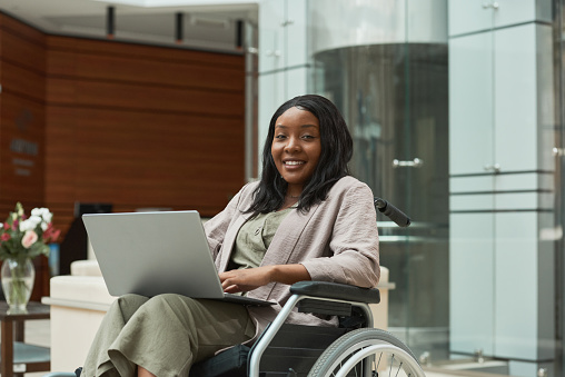Portrait of African disabled woman smiling at camera while sitting in wheelchair and working on laptop