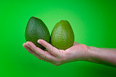Human hand holding two whole avocado fruits on green background.