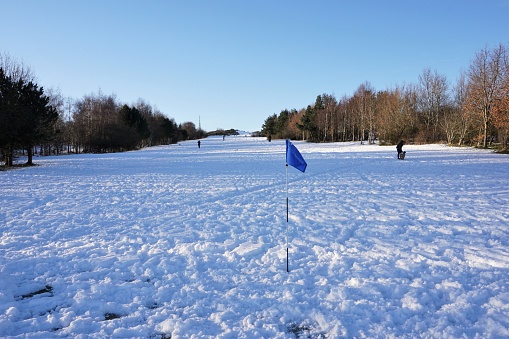 Edinburgh, Scotland - January 24 2021: A golf flag marks the putting surface after a snowfall has covered this golf course in Edinburgh. Recreational users enjoying the snow have replaced golfers.