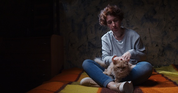 Distressed woman is sitting on the sofa in dark room and stroking a cat