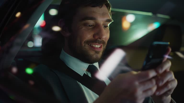 Happy Handsome Businessman in a Suit Commuting from Office in a Backseat of His Car at Night. Entrepreneur Using Smartphone while in Transfer Taxi in Urban City Street with Working Neon Signs.