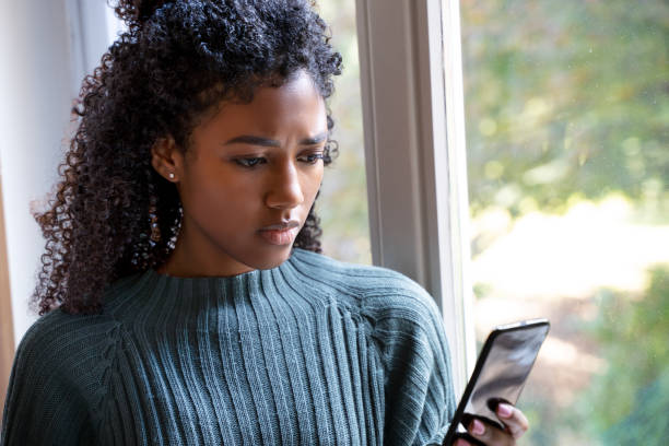 la femme noire regarde à l’extérieur de sa fenêtre regardant le portable. - suivre photos et images de collection