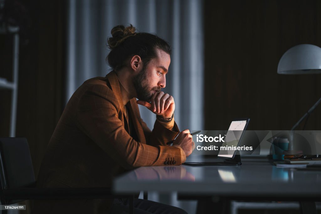 Pensive man working on laptop in office Side view of serious thoughtful adult bearded male in stylish wear sitting at table with modern laptop and analyzing business information while working in office in late evening Men Stock Photo
